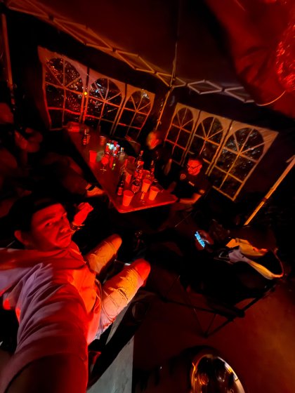 In einem dunkel und rot beleuchteten Zelt sitzen mehrere Personen an einem Tisch mit zahlreichen Flaschen und Bechern, während eine Person eine Selfieaufnahme macht.