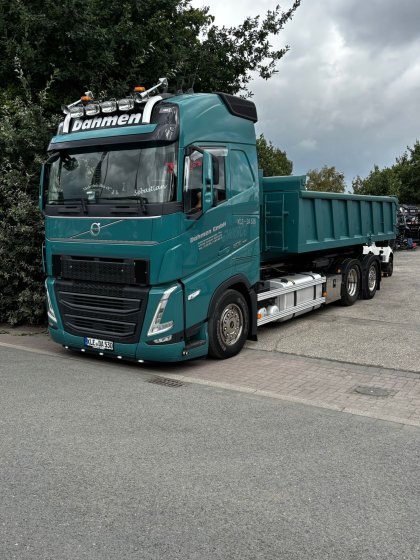 Ein grüner Volvo LKW mit Kipplaster-Anhänger der Firma Dahmen steht an einer Straße vor Bäumen unter bewölktem Himmel.
