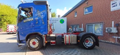 Blauer Lkw mit Aufschrift „Rippchen“ und tanzendem Skelett auf der Fahrertür, parkend vor einem Ziegelgebäude, daneben drei stehende Personen.