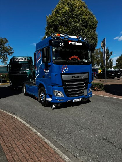 Ein blauer DAF-Sattelzug mit der Aufschrift "Fendrich" fährt auf einer asphaltierten Straße unter blauem Himmel, im Hintergrund ist ein grüner Lkw und eine große Baumkrone sichtbar.
