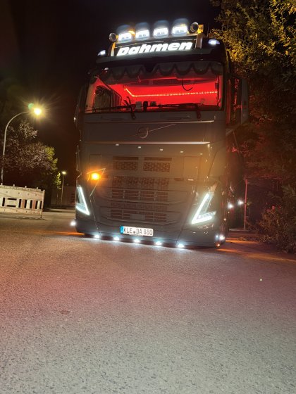Frontansicht eines beleuchteten grauen Volvo-Lastwagens mit „Dahmen“-Schriftzug bei Nacht auf einer Straße, umgeben von Bäumen und Straßenlaternen.