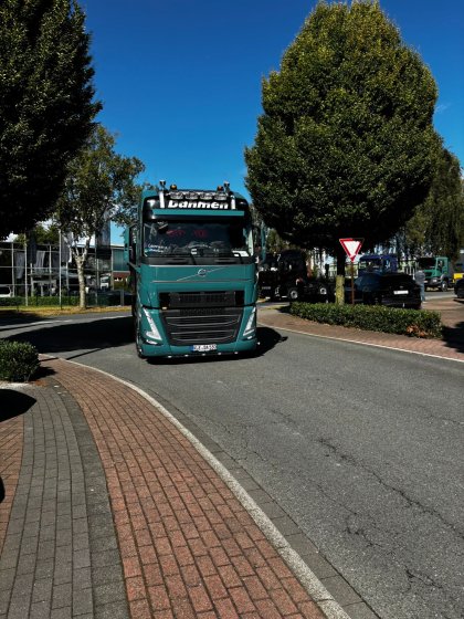 Grüner LKW mit dem Schriftzug "Dahmen" fährt auf einer Straße in einem Industriegebiet bei sonnigem Himmel, umgeben von mehreren Bäumen, einem Autoverkehrsschild und geparkten Fahrzeugen.