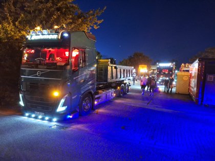 Bei Nacht sind mehrere LKWs mit leuchtenden Scheinwerfern auf einem belebten Parkplatz abgestellt, wo Menschen zwischen den Fahrzeugen gehen.