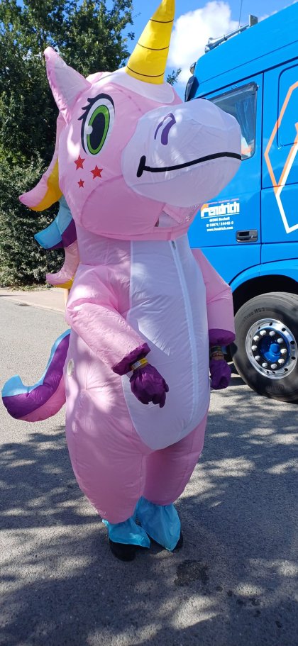 Person trägt auf einer Straße bei Sonnenschein ein aufblasbares pinkes Einhorn-Kostüm mit gelbem Horn, lila und blauen Details, vor einem blauen Lkw.