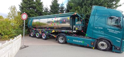 Blauer Volvo-LKW mit silbernem Tankauflieger, der mit bunten Wimpelgirlanden geschmückt ist und vor einem Verbotsschild auf einem gepflasterten Weg steht.