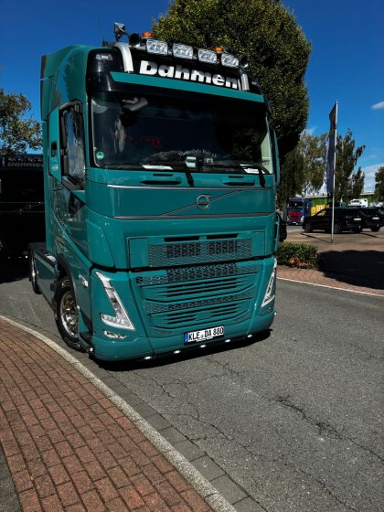 Grüner Volvo-LKW mit dem Schriftzug „Dahmen“ auf der Windschutzscheibe steht auf einem Parkplatz neben einem Gehweg aus roten Pflastersteinen unter blauem Himmel.