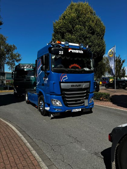 Blauer DAF-Sattelzug auf einer Straße in einem Industriegebiet bei klarem Himmel, dahinter grüner LKW und rechts eine Fahne auf einem Gehweg.
